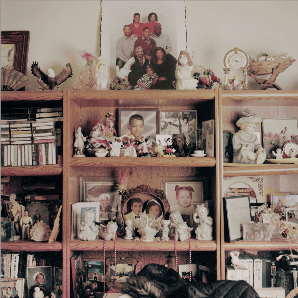 A family bookshelf with photos and figurines. Some objects include rosaries, cassette tapes, books, small clocks, and a teacup.